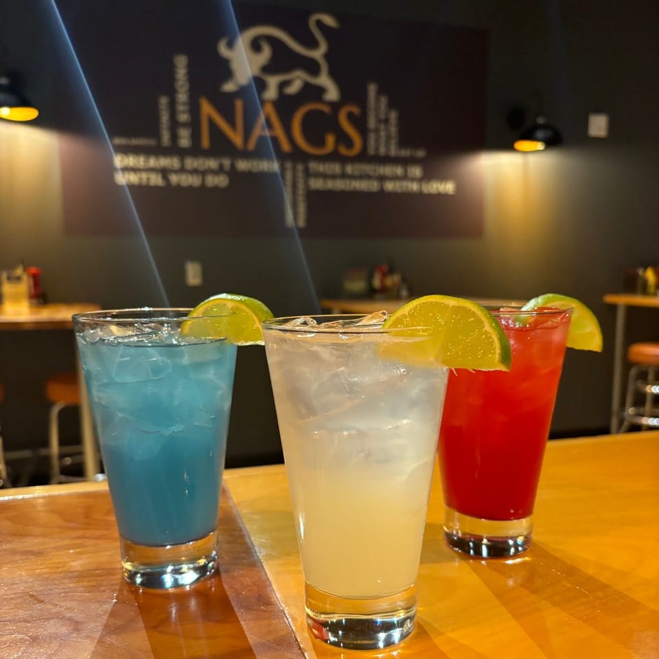 Three colorful cocktails with lime wedges on a wooden bar, with the NAGS logo in the background.