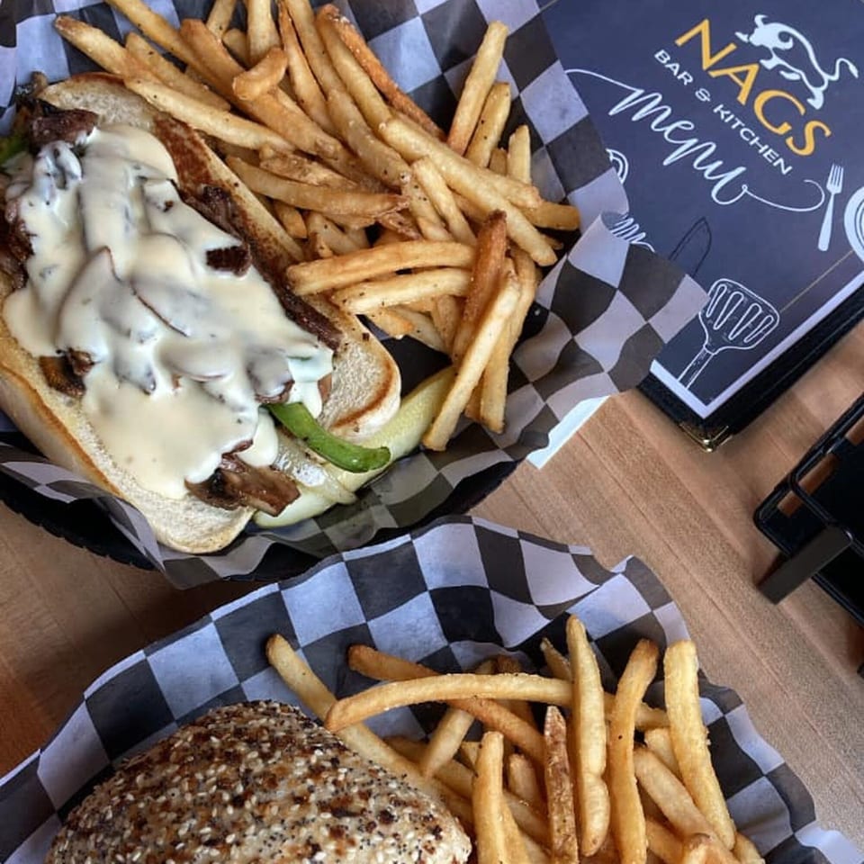 Cheesesteak with fries and a burger with fries, served in checkered paper-lined baskets next to a NAGS menu.
