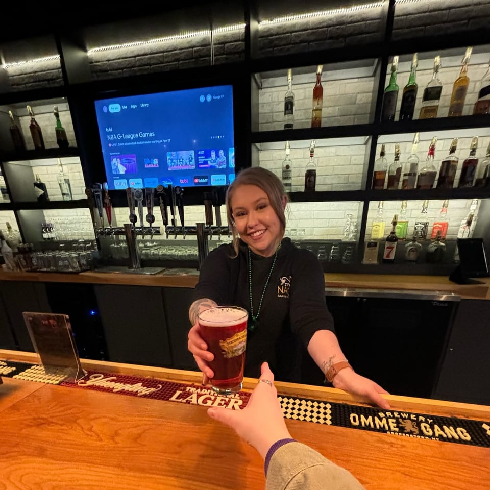 A smiling NAGS Bar and Kitchen bartender serves a beer to a customer across a wooden bar.